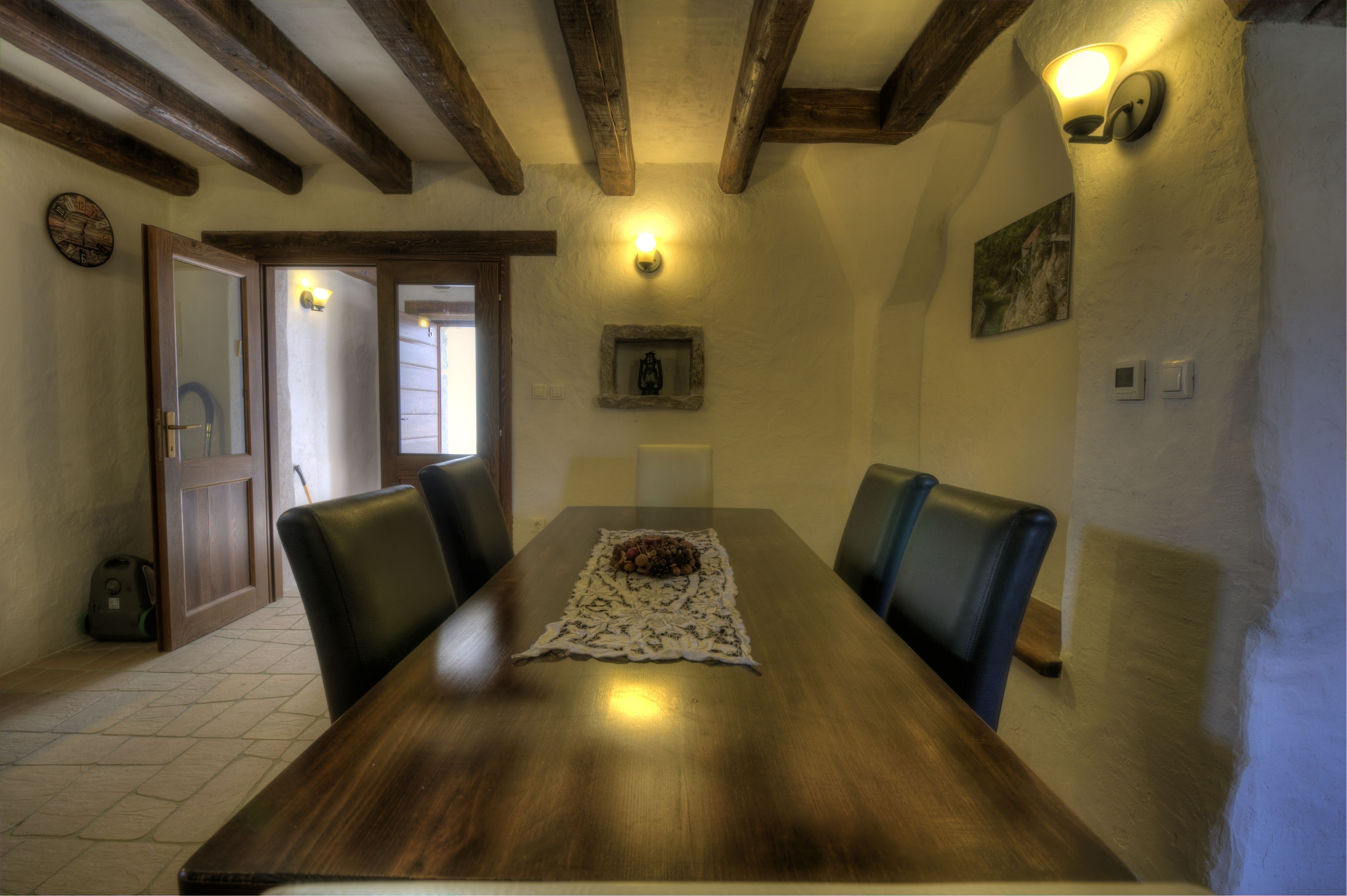 A wooden dining table with a lace table runner and a bowl of fruit. There are four chairs around the table. The room has exposed wooden beams on the ceiling and a wall-mounted light fixture.