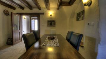 A wooden dining table with a lace table runner and a bowl of fruit. There are four chairs around the table. The room has exposed wooden beams on the ceiling and a wall-mounted light fixture.