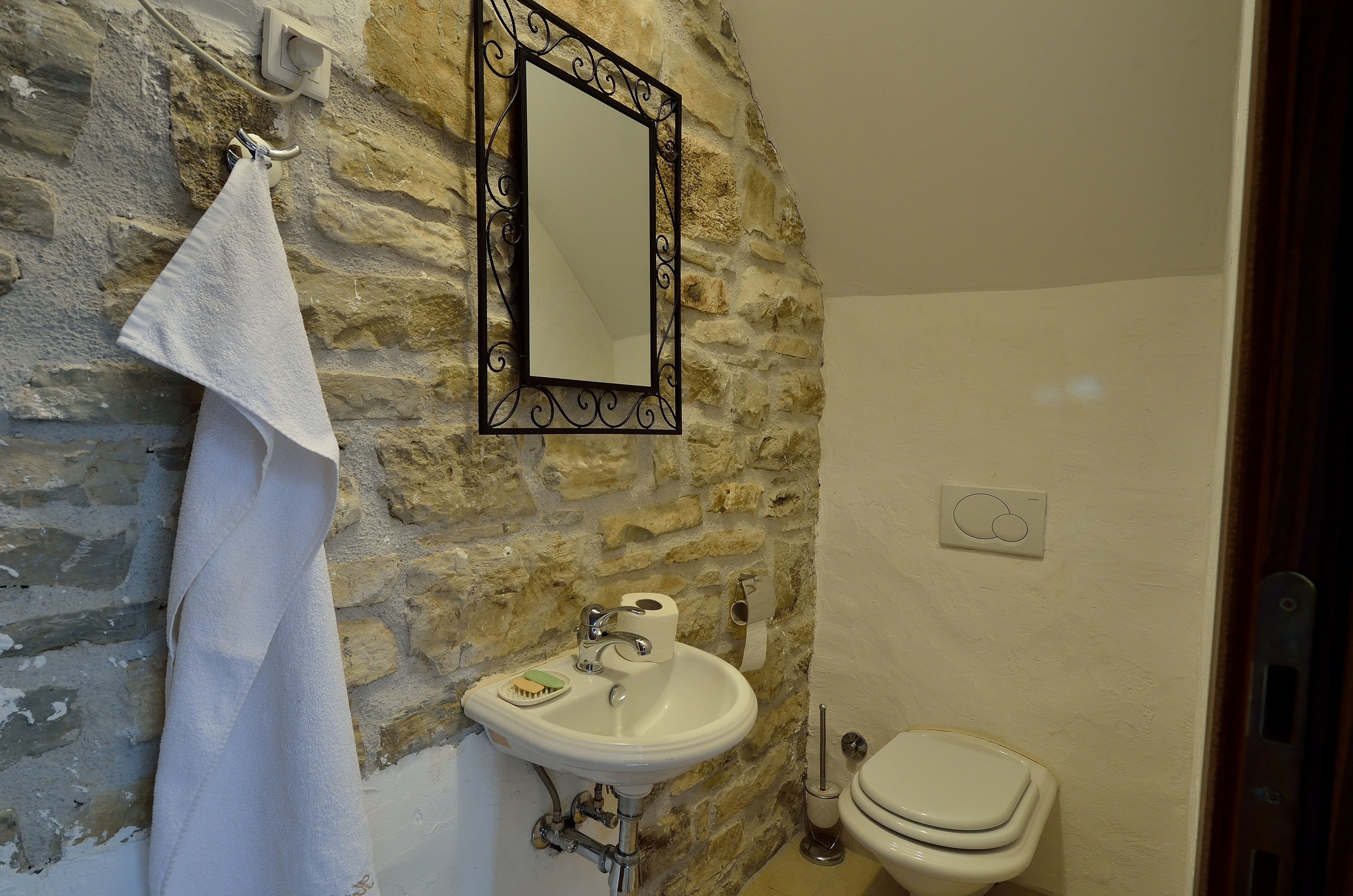 A bathroom with a stone wall, a white sink, a mirror, a towel, a toilet, and a soap dish.