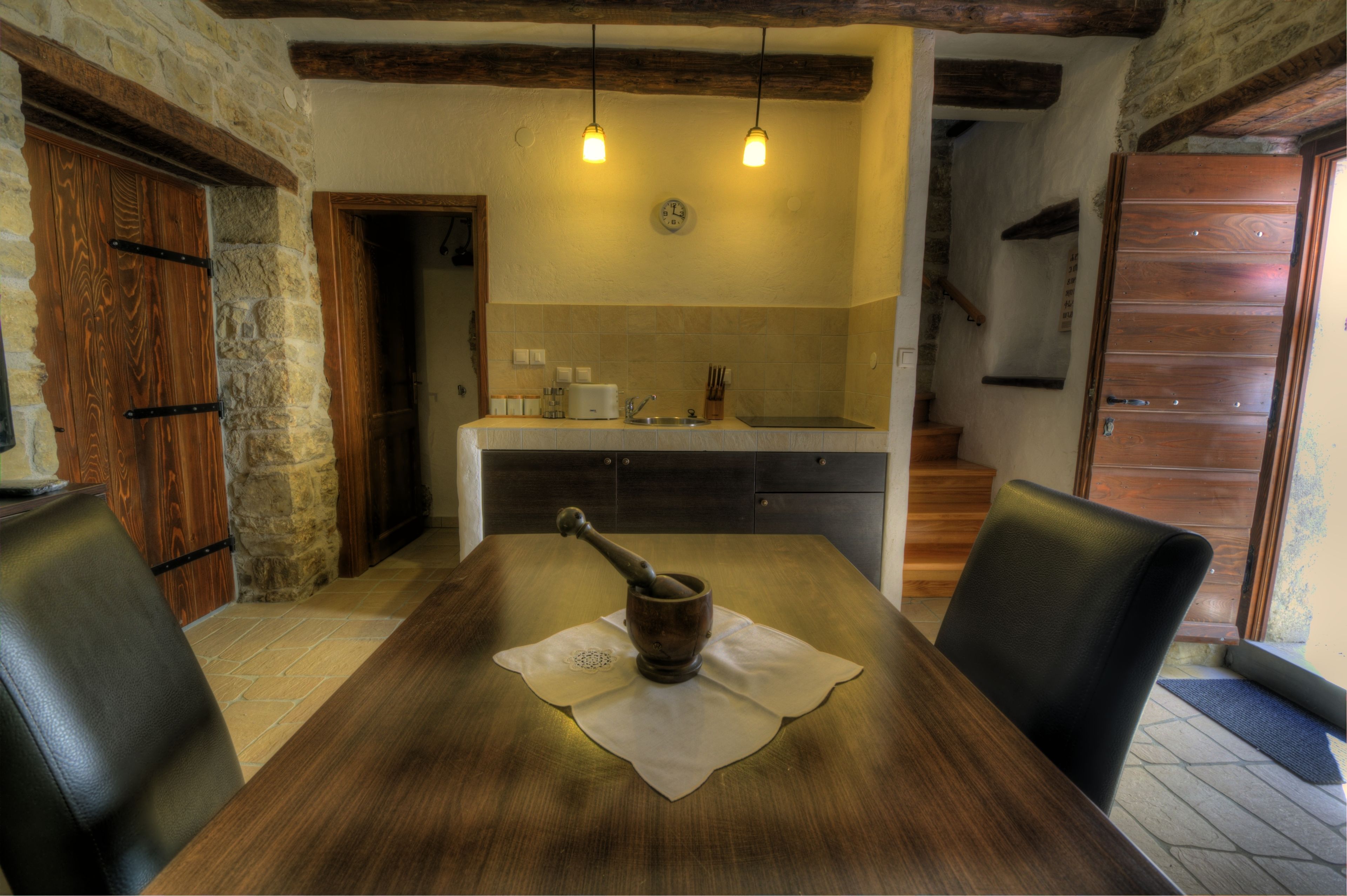 Une table à manger en bois, agrémentée d’un mortier et d’un pilon rustiques en guise de décoration centrale, un mur en pierre et une cuisine en arrière-plan.