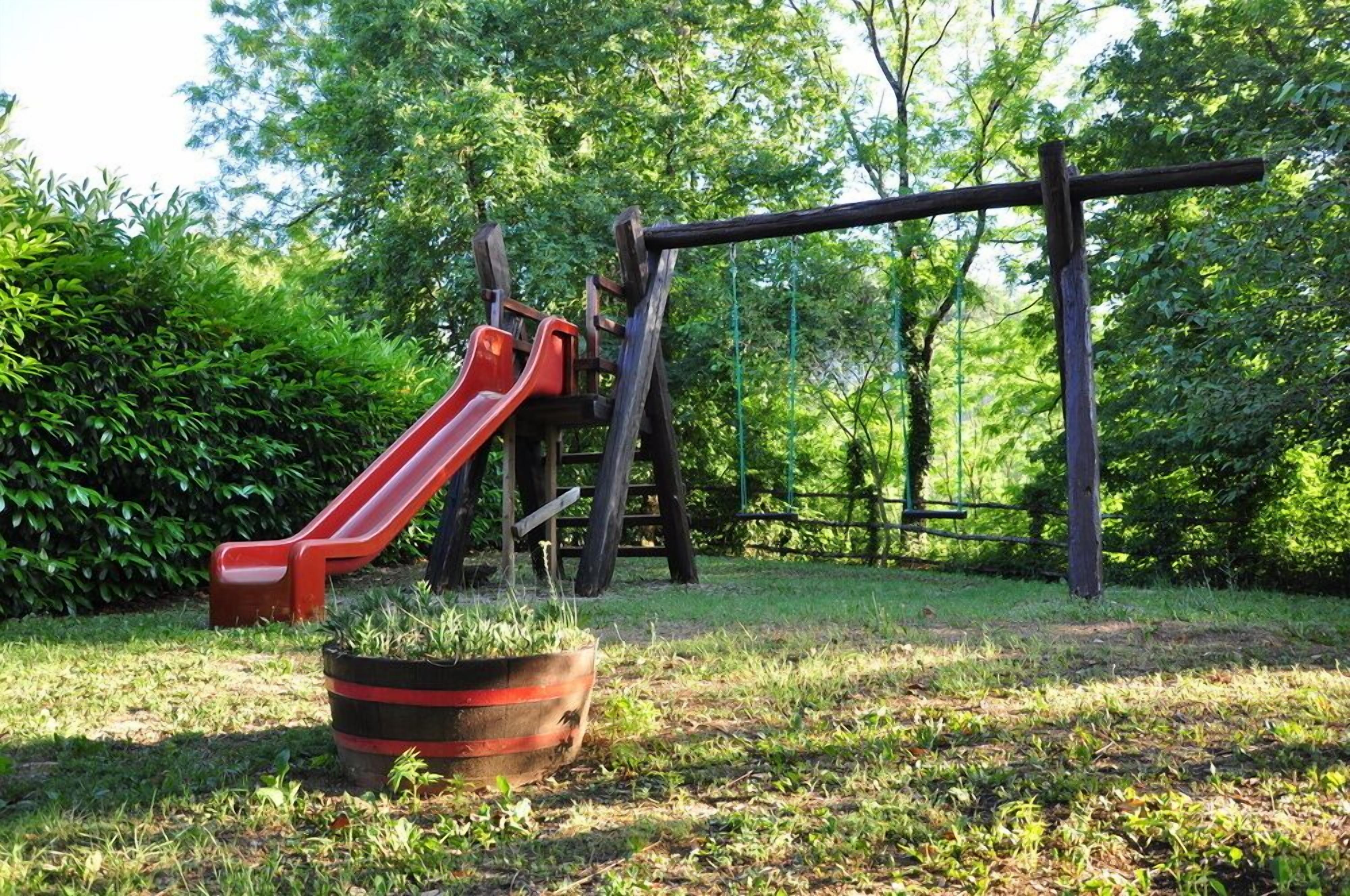 A playground with a red slide and a wooden swing set in a grassy area surrounded by trees.