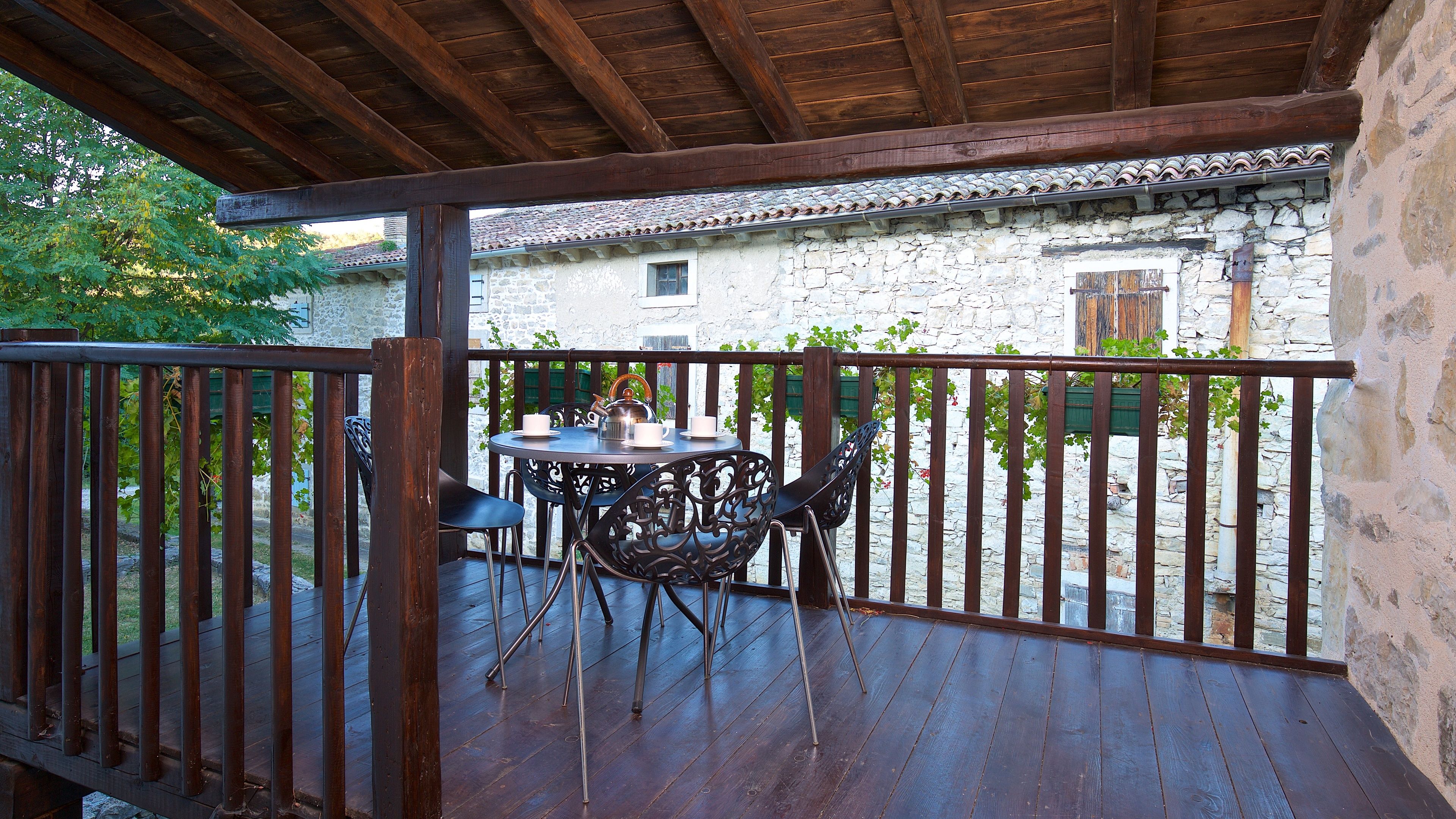 Eine Holzterrasse mit Tisch und Stühlen, im Hintergrund ein Steinhaus und ein Geländer aus Holz.