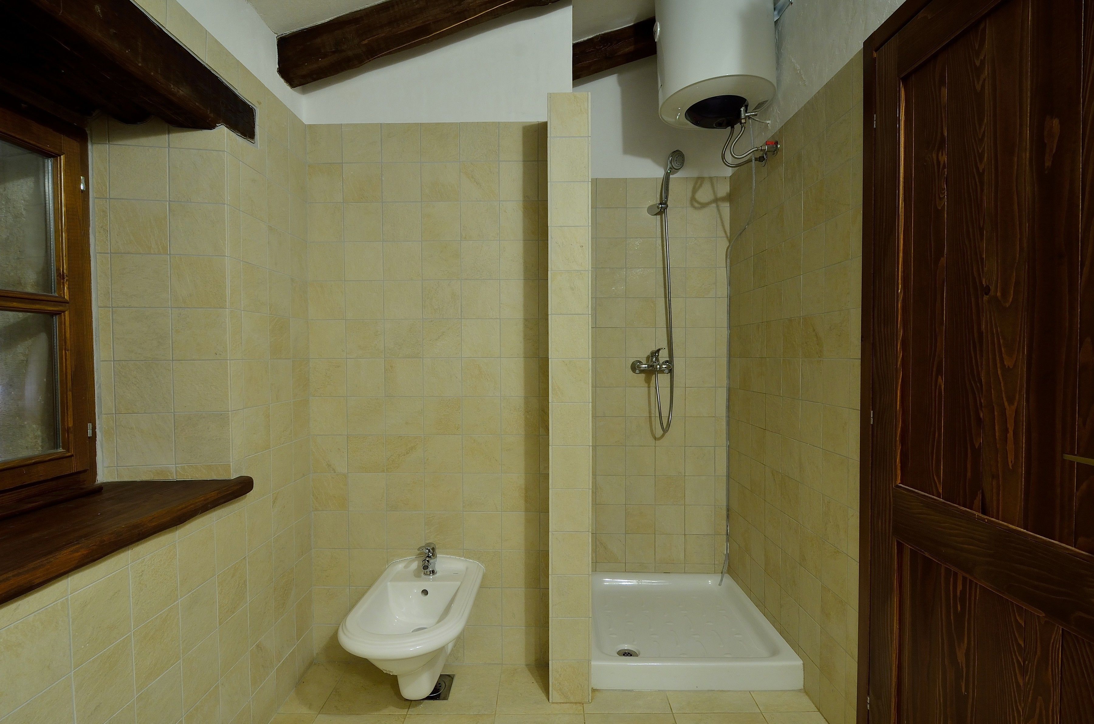 A bathroom with beige tiles, a wooden vanity, a bidet, a shower, and a wooden door.