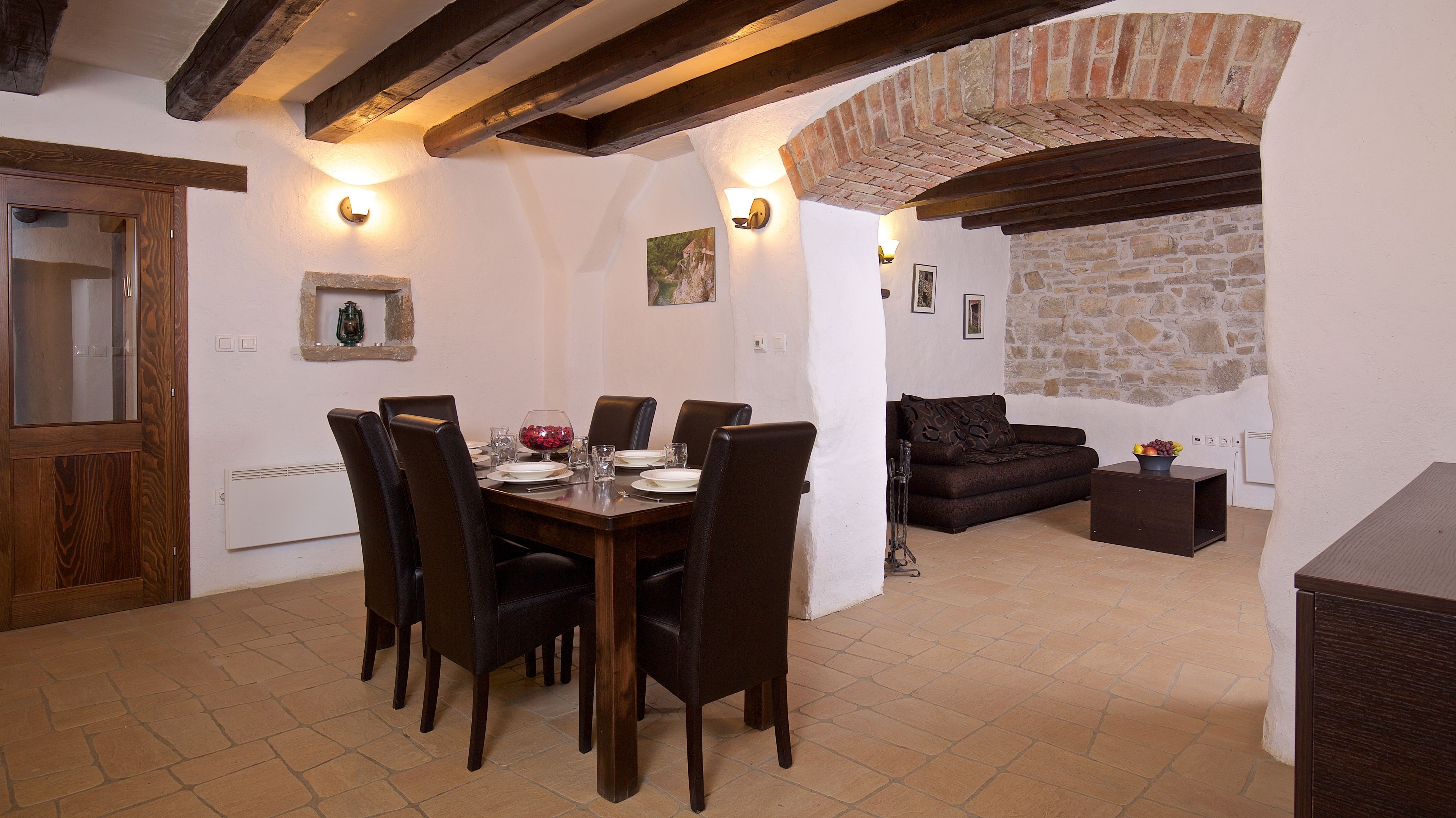 A dining area with a wooden table set for six, surrounded by leather chairs. There is a stone wall and a brick archway in the background.