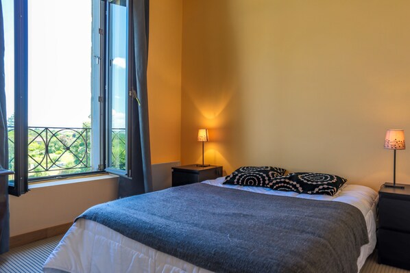 A bedroom with a bed, two bedside tables, a window with curtains, and a view of greenery outside.