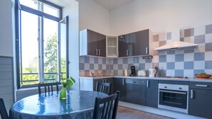 A modern kitchen with grey cabinets, a built-in oven, and a dining area with a round table and chairs. - Résidence du Chateau de Bégué (Cazaubon)
