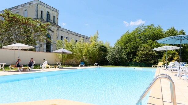 A swimming pool with lounge chairs and umbrellas, surrounded by greenery and a building in the background. - Résidence du Chateau de Bégué (Cazaubon)