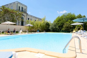 A swimming pool with lounge chairs and umbrellas, surrounded by greenery and a building in the background.