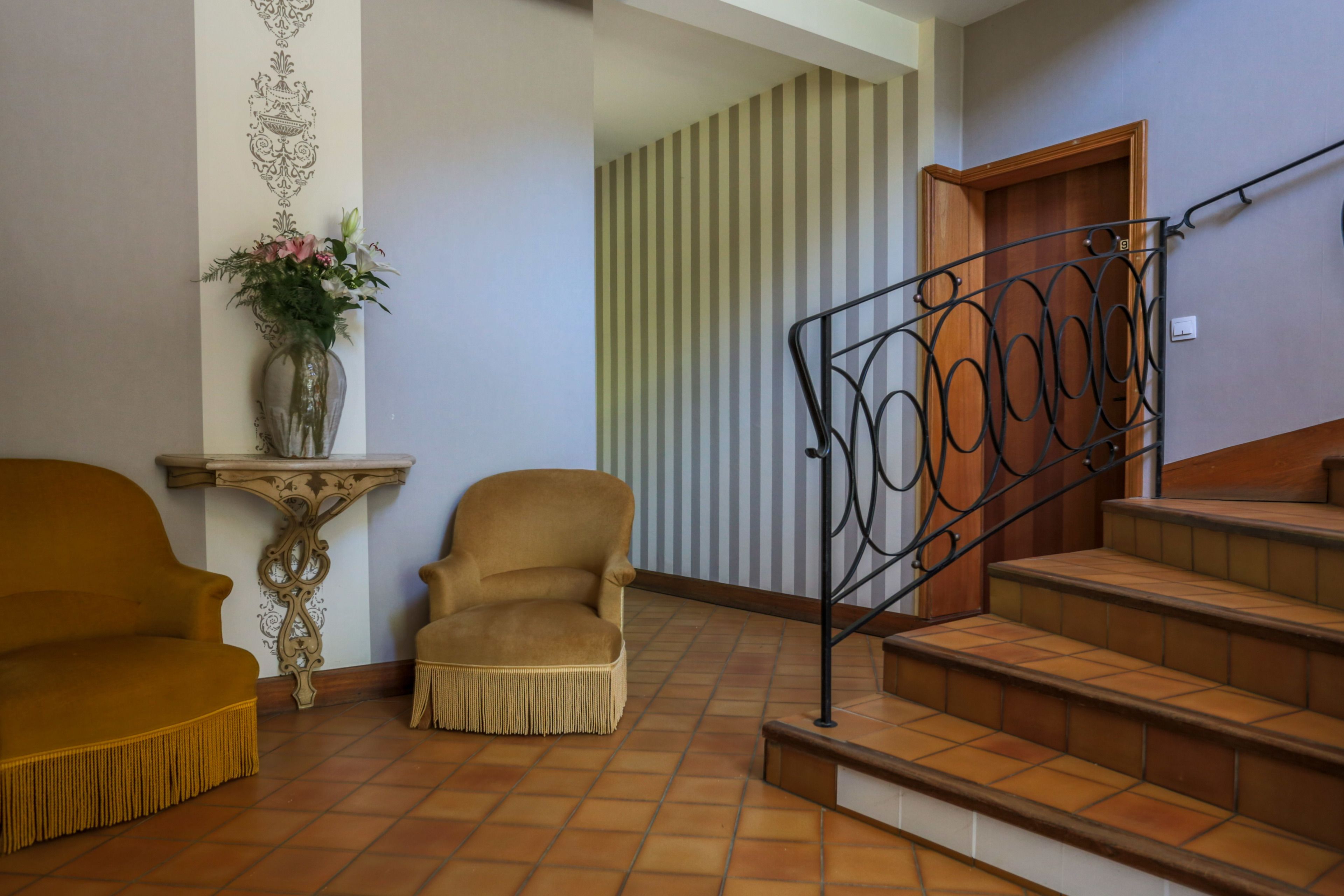 A staircase with a wrought iron railing, a brown leather sofa, a beige armchair, and a decorative vase with flowers on a small table.