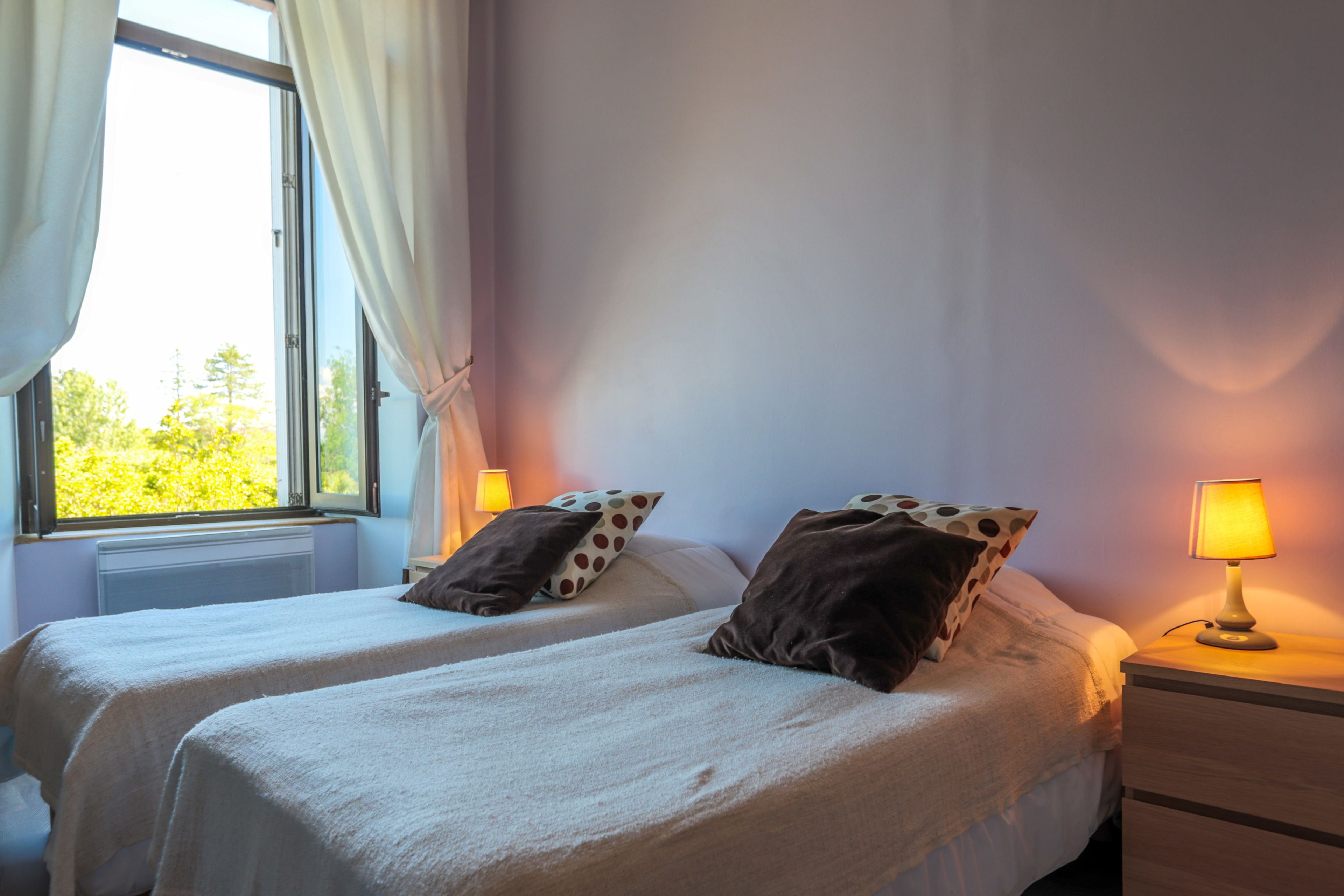 Un dormitorio con dos camas, una ventana con cortinas, una mesita de noche con lámpara y vistas a árboles.