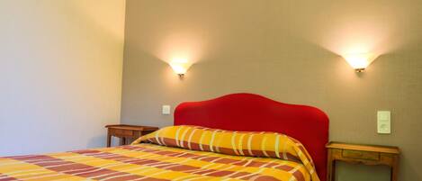 Habitación de hotel con cabecera roja, colcha a cuadros, dos lámparas de noche y una mesita de noche.