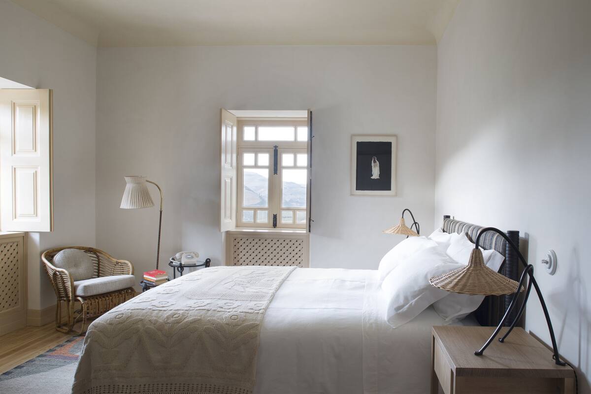 Une chambre avec un grand lit, une chaise en osier, une table de chevet avec une lampe et une fenêtre donnant sur un beau paysage.