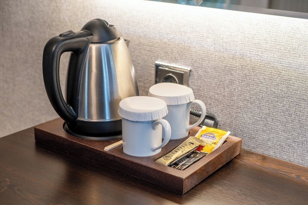 A coffee maker, two mugs, and a tray with coffee packets on a wooden surface.