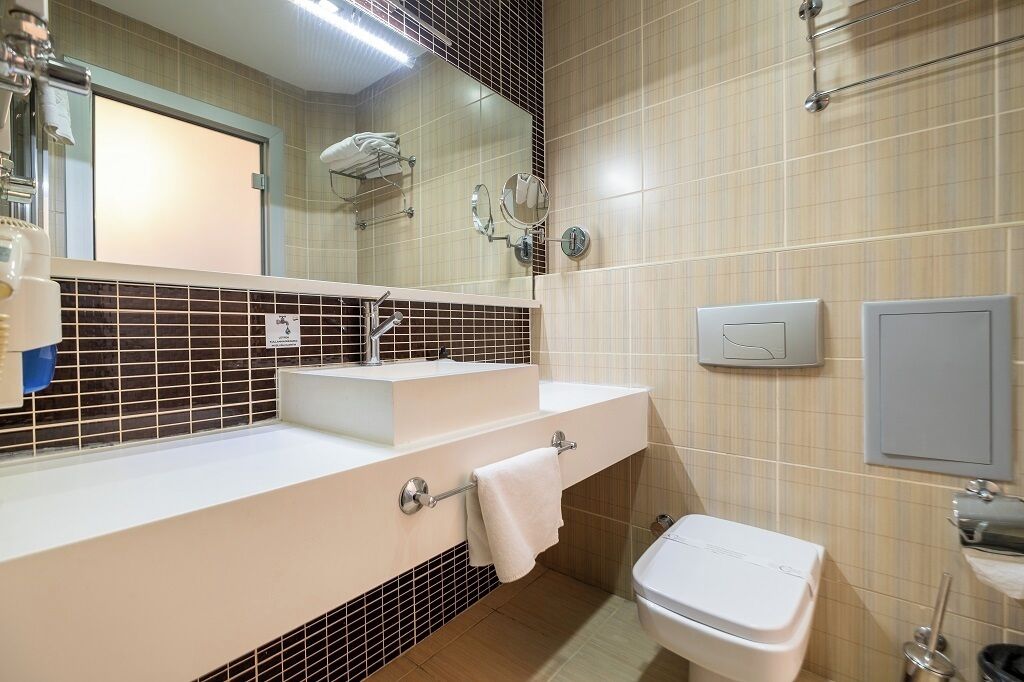 A bathroom with a sink, mirror, towel rack, toilet, and a wall-mounted light switch.