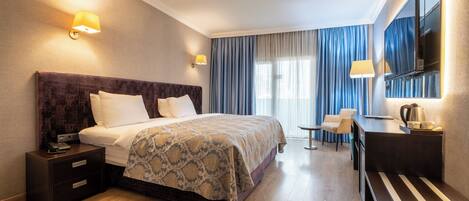 A hotel room with a large bed, bedside tables, a desk with a chair, a mirror, and a window with curtains.