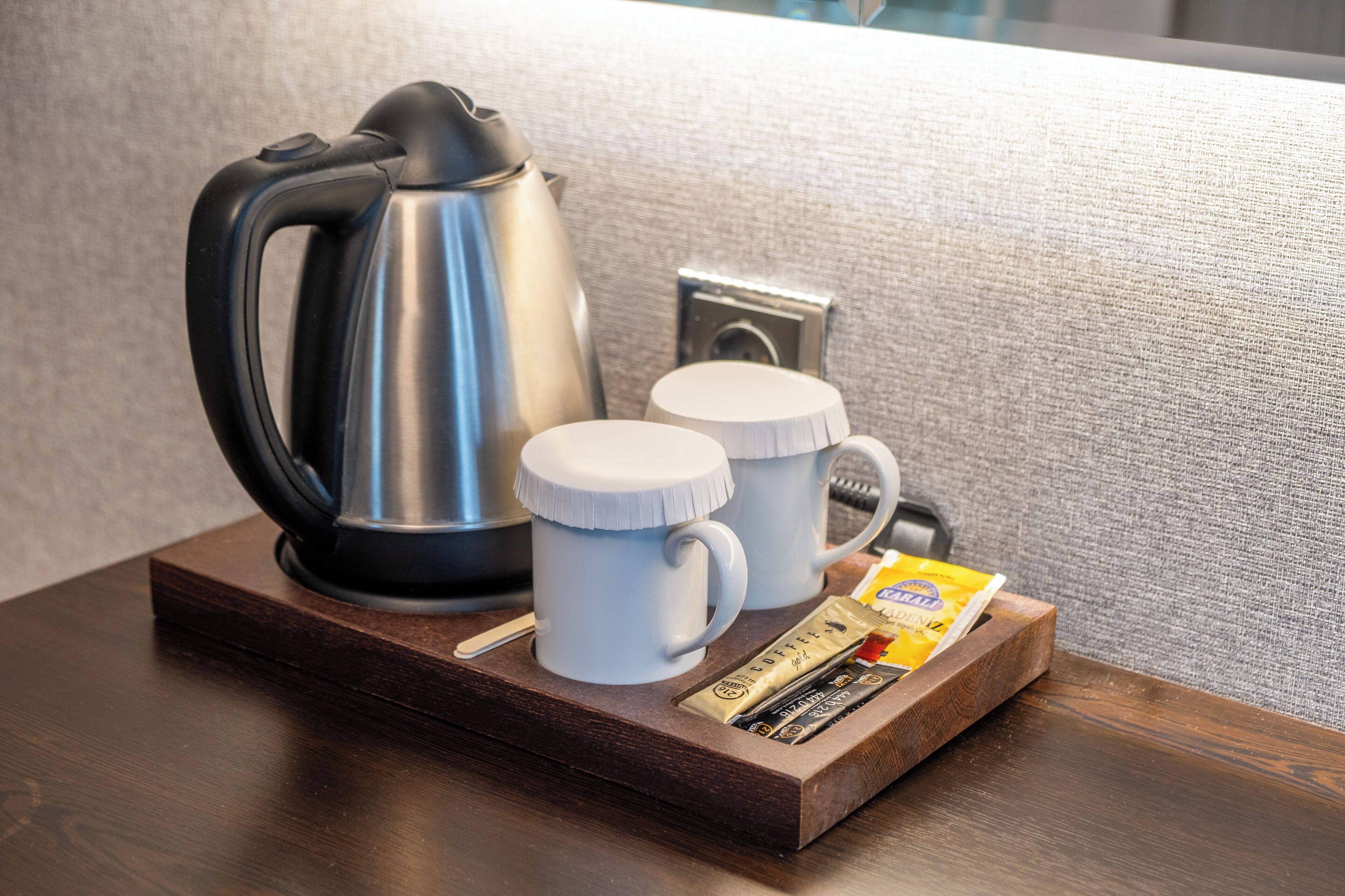 A wooden tray with a stainless steel kettle, two white mugs, and a packet of coffee creamer on a wooden table.