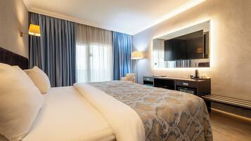 Habitación de hotel con una cama grande, un escritorio con televisor de pantalla plana, una lámpara y un ventanal con cortinas.