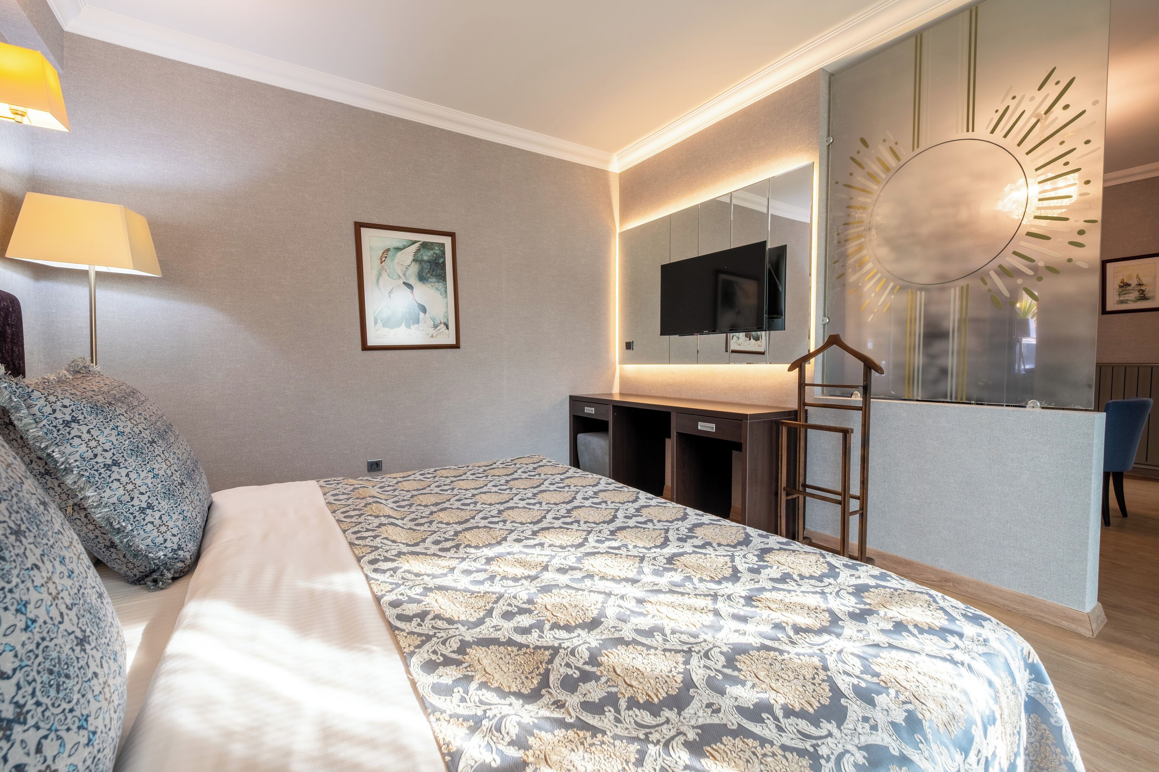 A hotel room with a bed, bedside lamp, a desk with a mirror, a TV, and a decorative wall piece.
