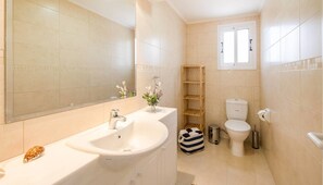 Un baño con lavamanos, espejo, inodoro y una pequeña ventana.
