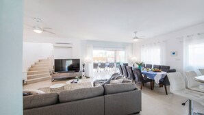 Un saló modern amb un sofà gris gran, un televisor de pantalla plana, una zona de menjador amb taula per a quatre i una escala que mena a un pis superior.