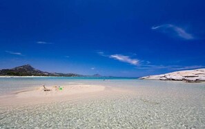 A beach with clear shallow waters, a sandy shore, and rocky formations in the background.
