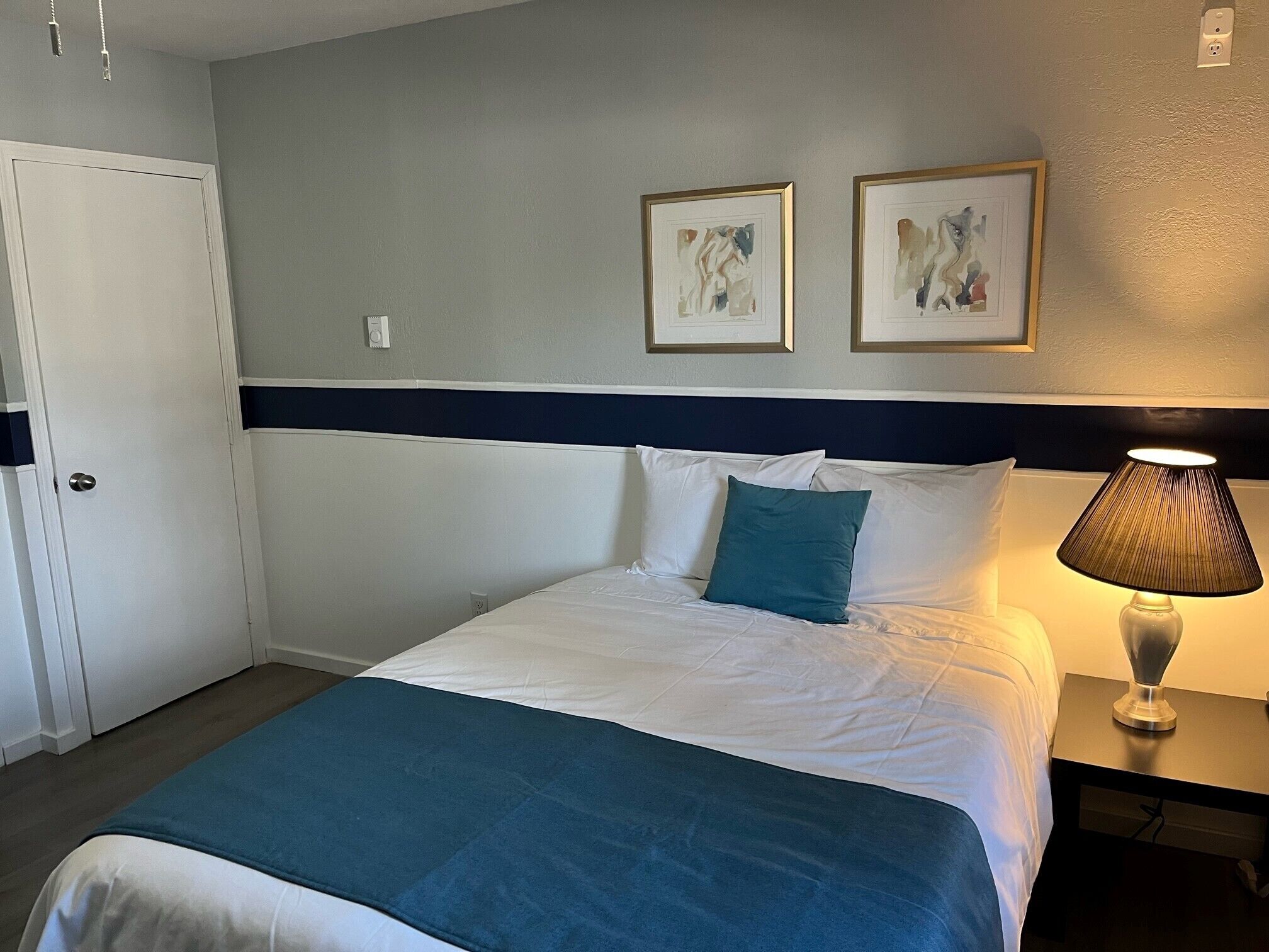 Quarto de hotel com cama, mesa de cabeceira com abajur e duas obras de arte emolduradas na parede.