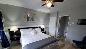 Un dormitorio con cama, dos mesitas de noche con lámparas, un ventilador de techo, un cuadro enmarcado en la pared y una puerta que da a otra habitación.