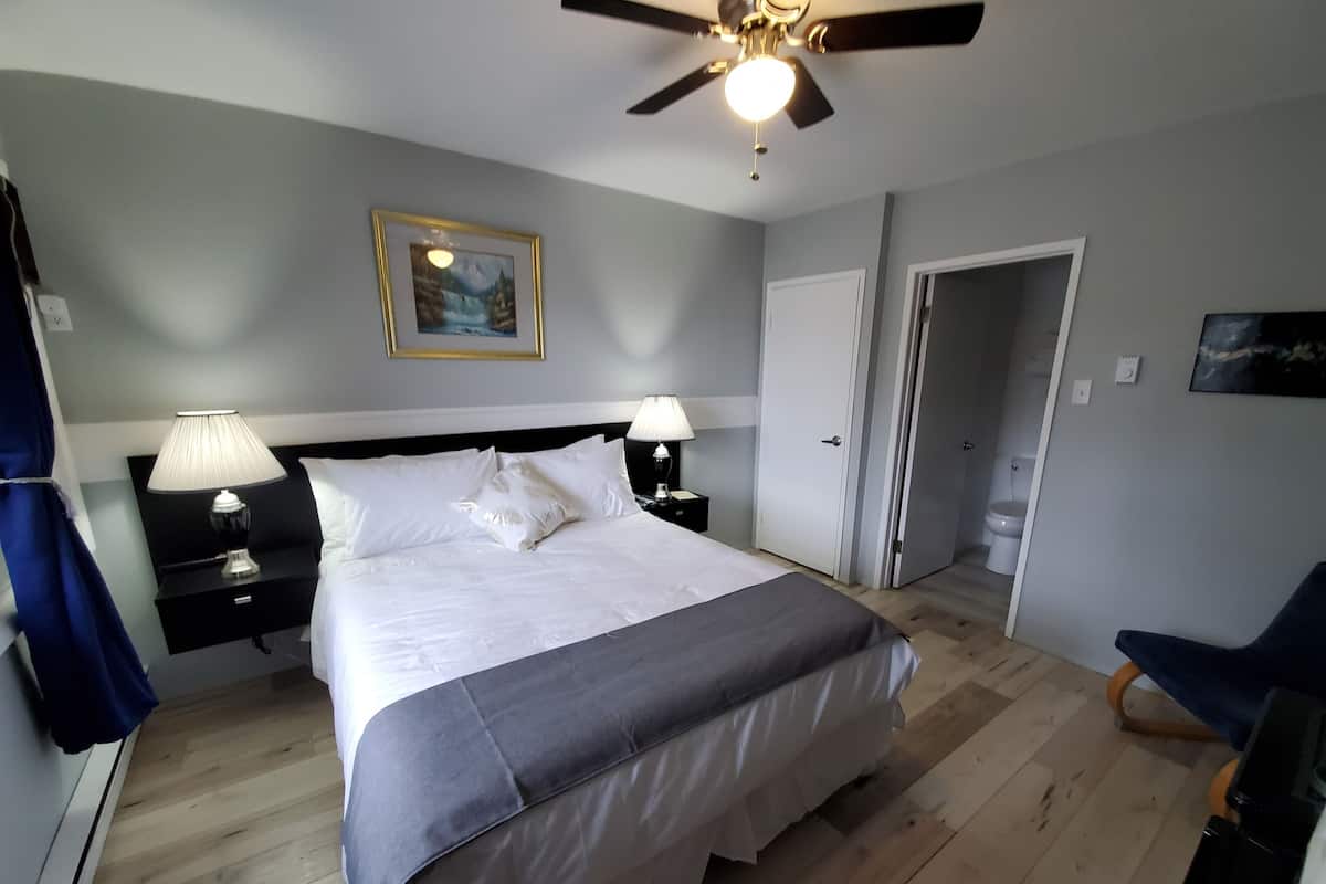 A bedroom with a bed, two bedside tables with lamps, a ceiling fan, a framed picture on the wall, and a door leading to another room.