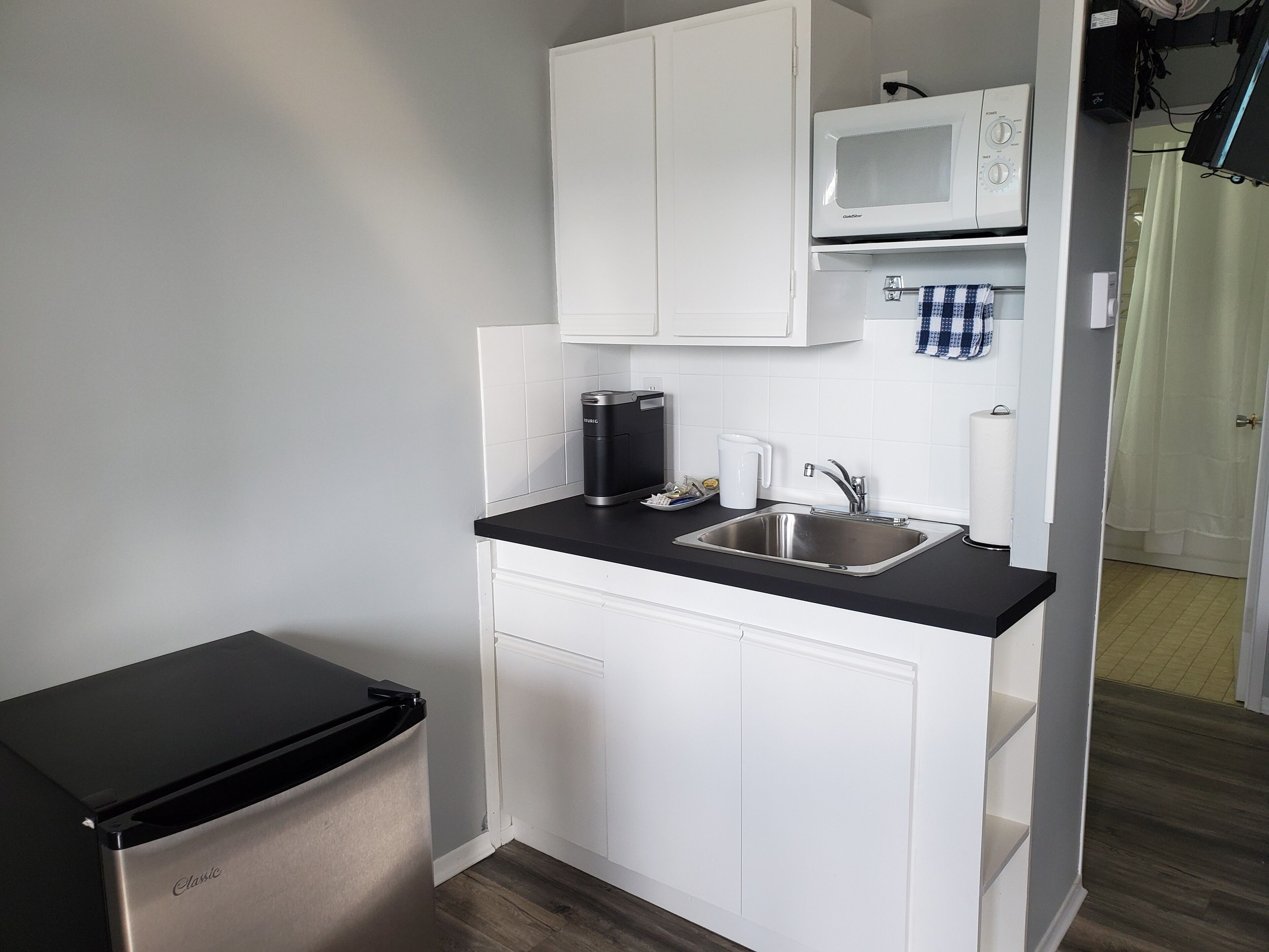 A compact kitchenette with a black countertop, stainless steel sink, white cabinets, a microwave, and a black refrigerator.