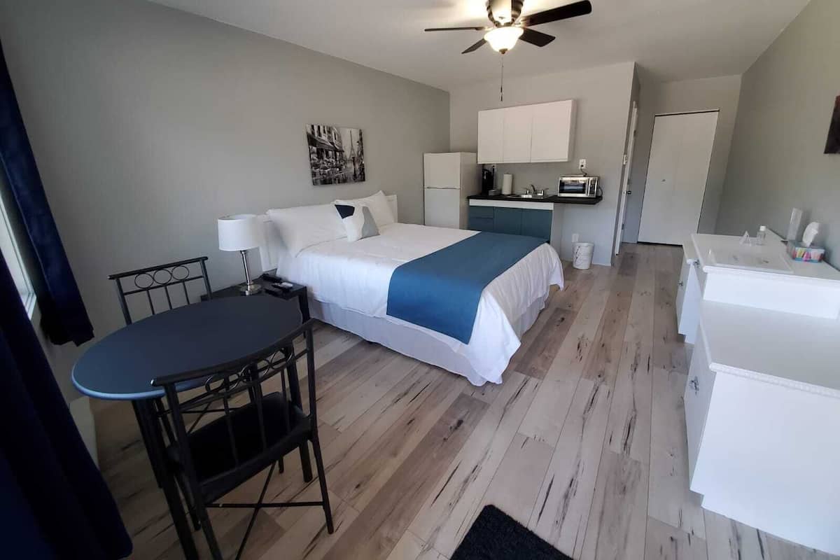 A bedroom with a bed, bedside table, desk, chair, kitchenette, and a ceiling fan.