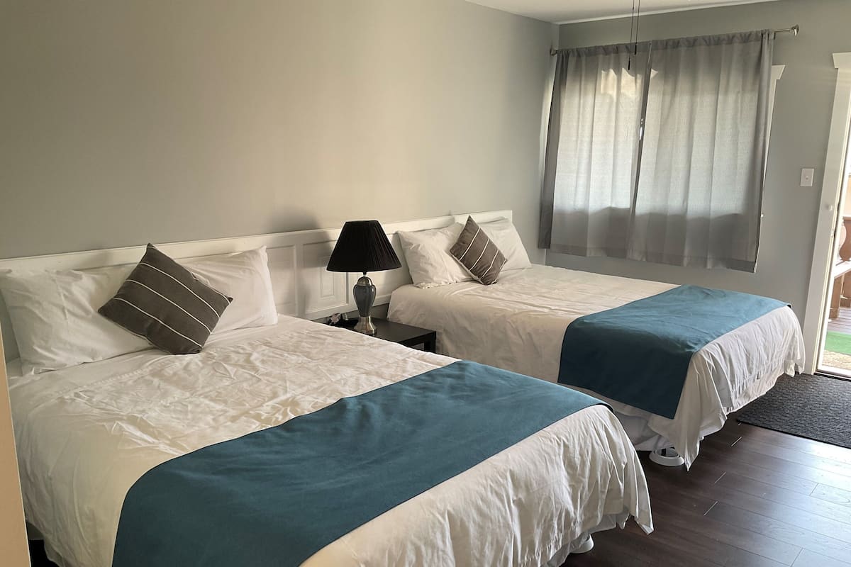 A hotel room with two beds, each with a white and blue bedspread, a nightstand with a lamp, and a window with curtains.