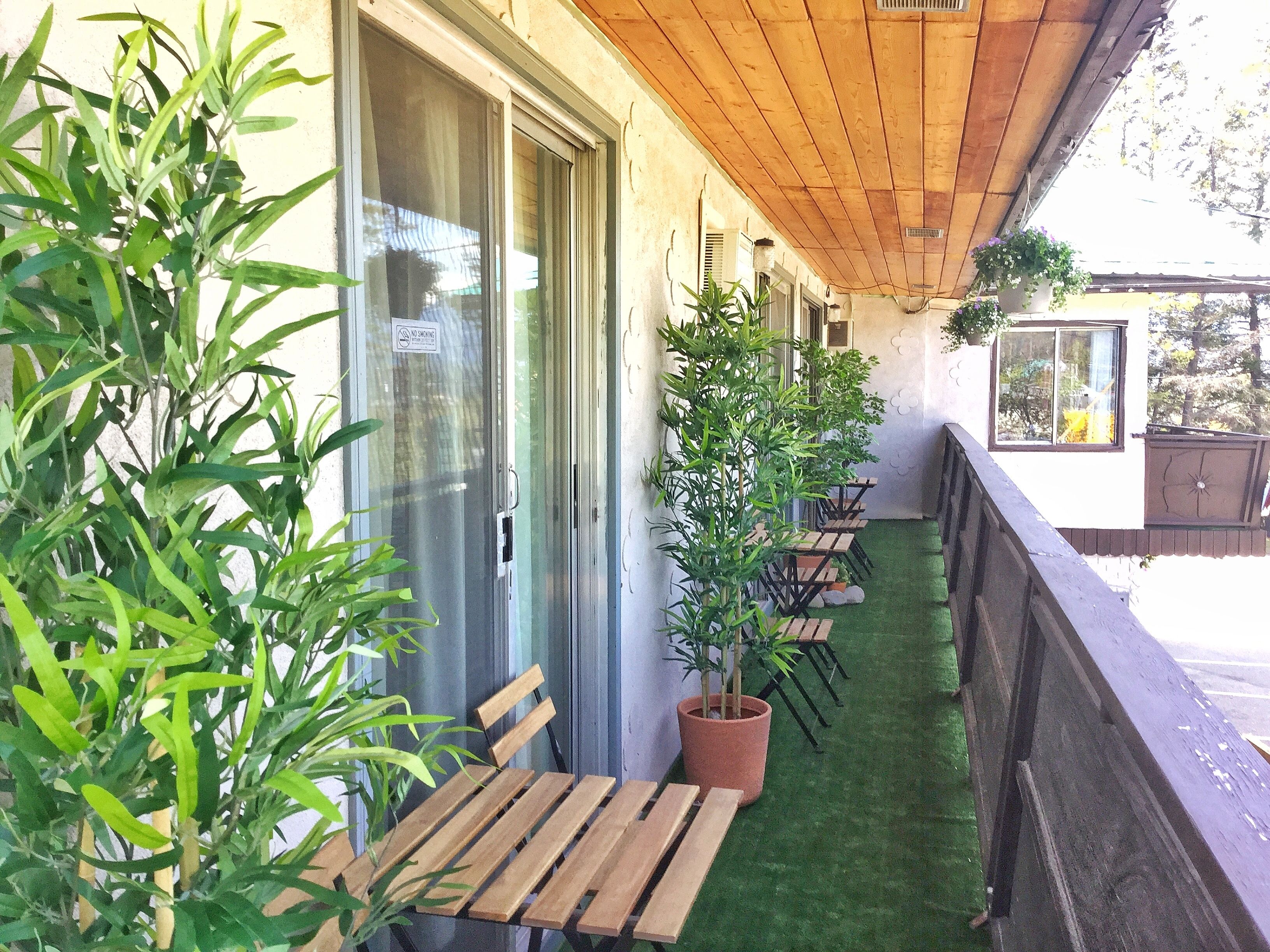 Un espace extérieur abrité, avec des tables et des chaises en bois, des plantes en pot et une porte en verre sur laquelle est apposé un panneau indiquant qu'il est interdit de fumer.