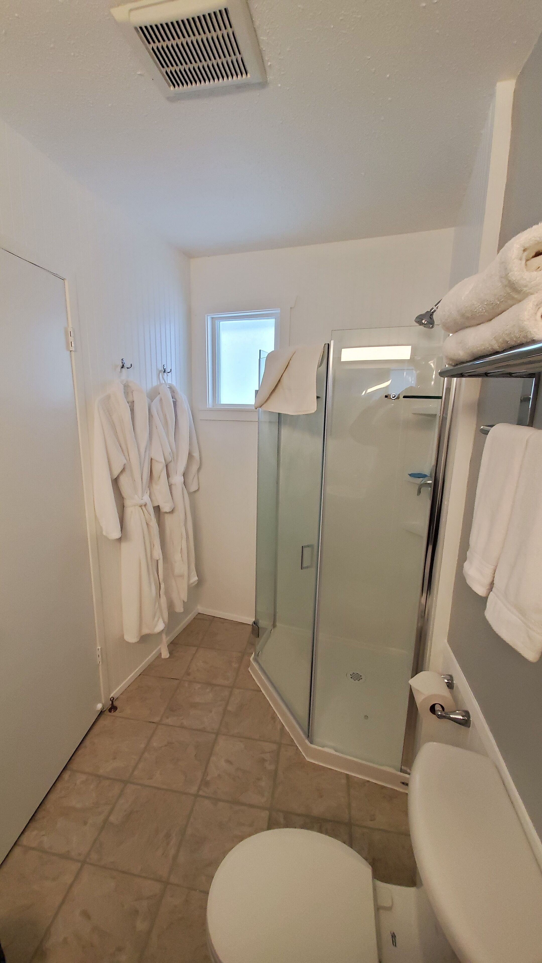 Une salle de bain avec une douche à parois de verre, des toilettes et un lavabo. Des serviettes et des peignoirs blancs sont suspendus à des crochets.