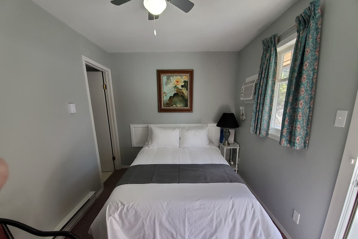 A neatly made bed with white bedding, a ceiling fan, a bedside lamp, and a framed painting on the wall.