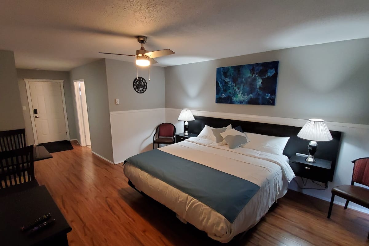 A neatly arranged hotel room with a large bed, two bedside tables with lamps, a ceiling fan, a wall clock, and a painting.