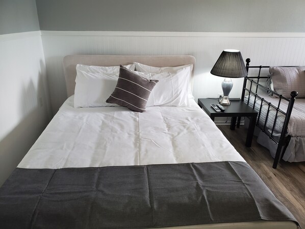 A neatly made bed with white and dark grey bedding, a bedside table with a lamp, and a chair with a cushion. - Celadon Lodge (Radium Hot Springs)