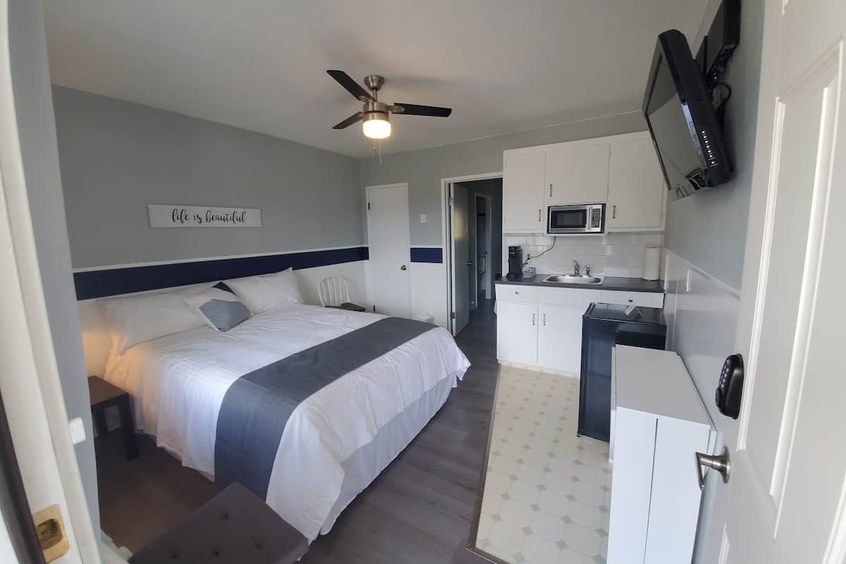 A compact hotel room with a bed, bedside table, and a small desk. A ceiling fan is visible, and a microwave is mounted above the kitchen area.