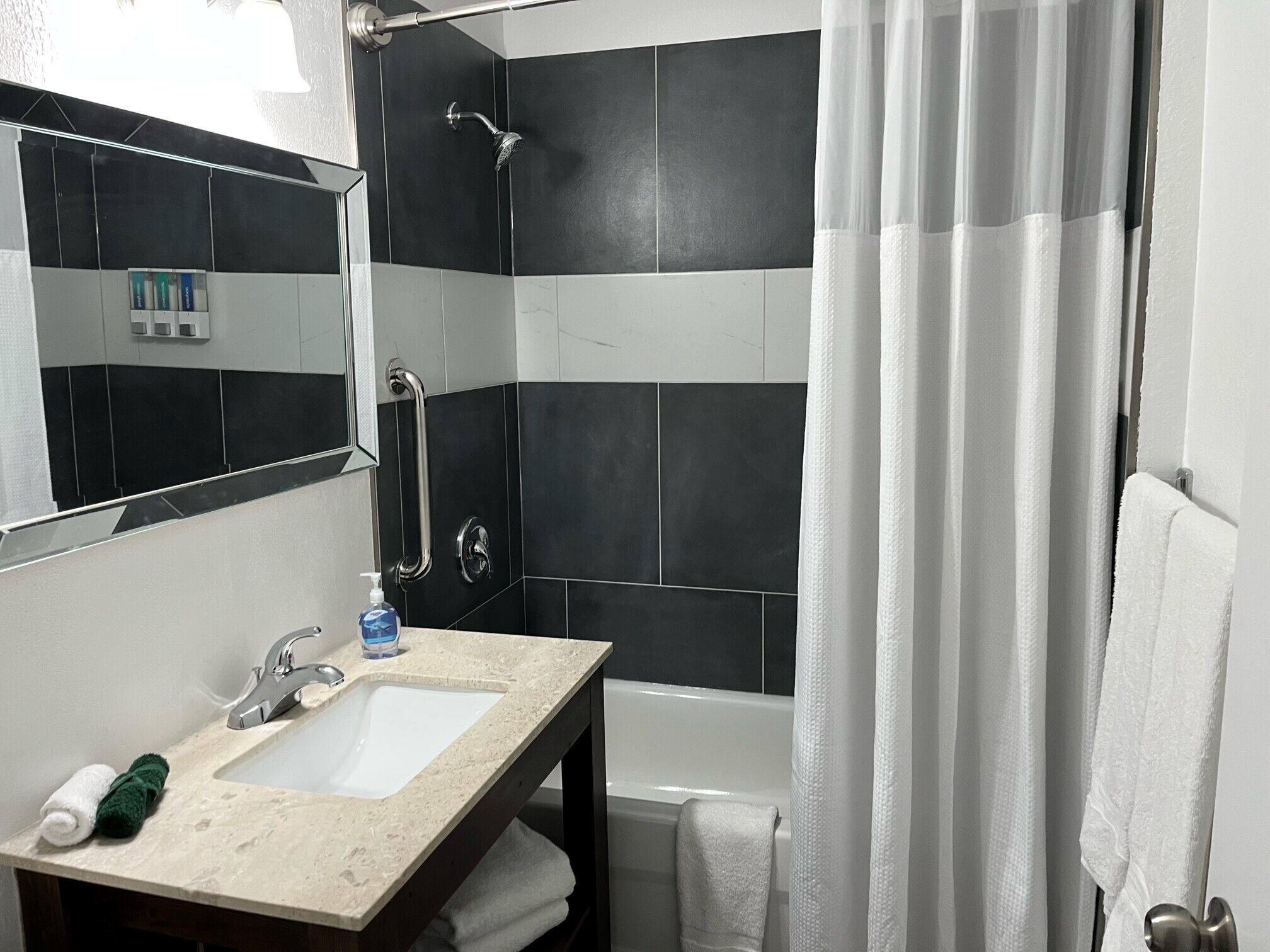 Un baño con lavamanos, espejo y ducha con barra de apoyo.