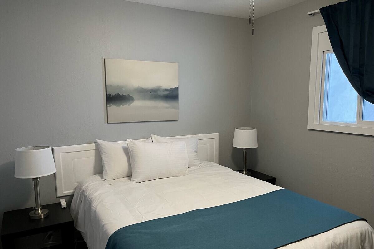 A neatly made bed with white bedding and a teal accent, two bedside lamps, a window with dark curtains, and a painting of a landscape on the wall.