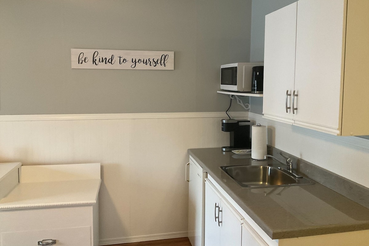 A small kitchenette with a microwave, coffee maker, and a sink. There is a sign on the wall that says "be kind to yourself."
