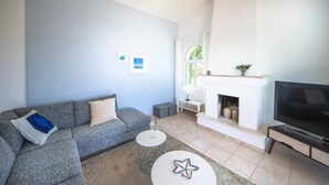 A modern living room with a grey sofa, a flat-screen TV, a fireplace, and a window with a view of greenery. - Villa Luna (Pegeia)
