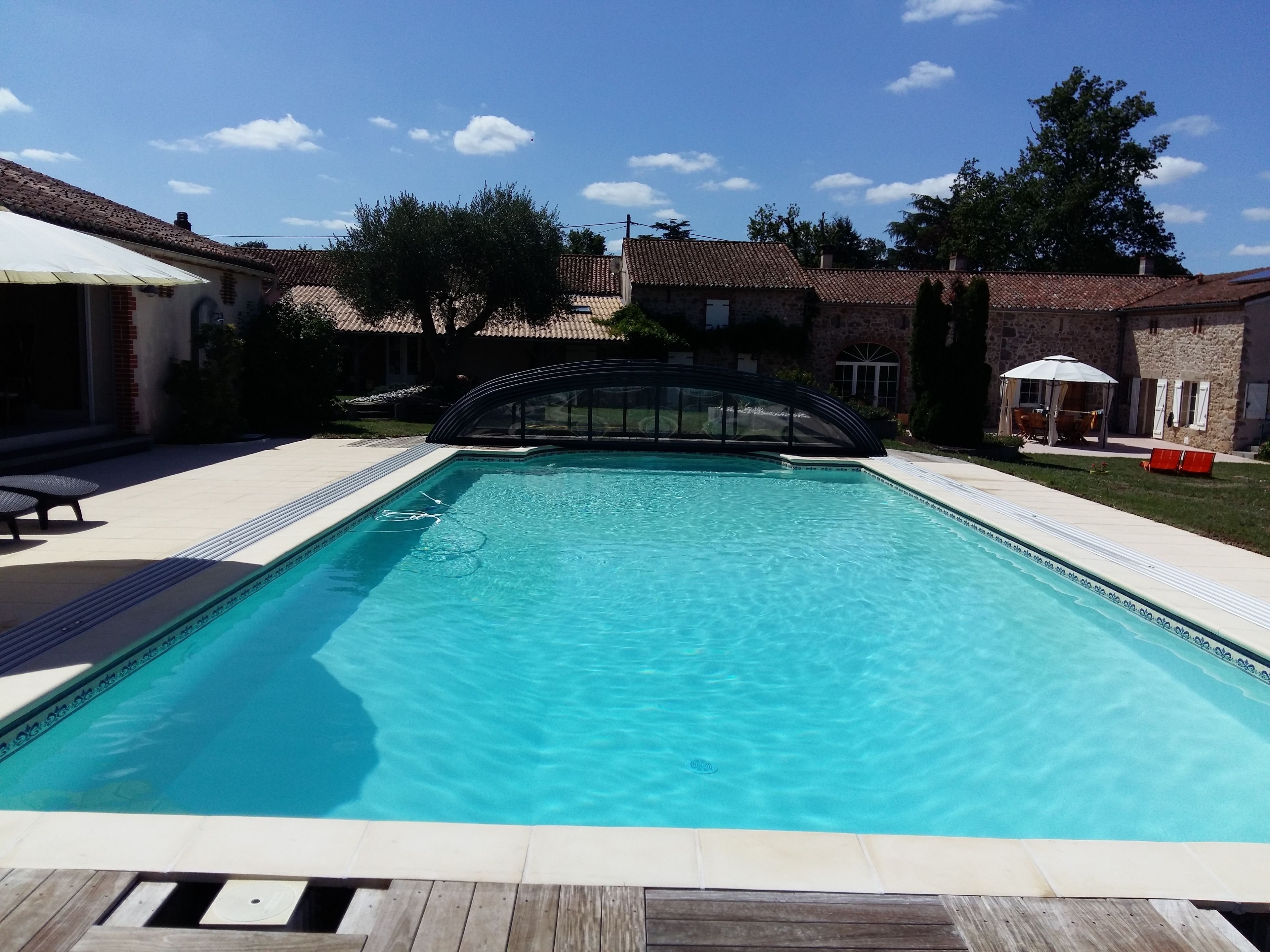 Une piscine rectangulaire avec une terrasse en bois au premier plan, entourée d’un bâtiment en pierre doté d’un toit en tuiles.