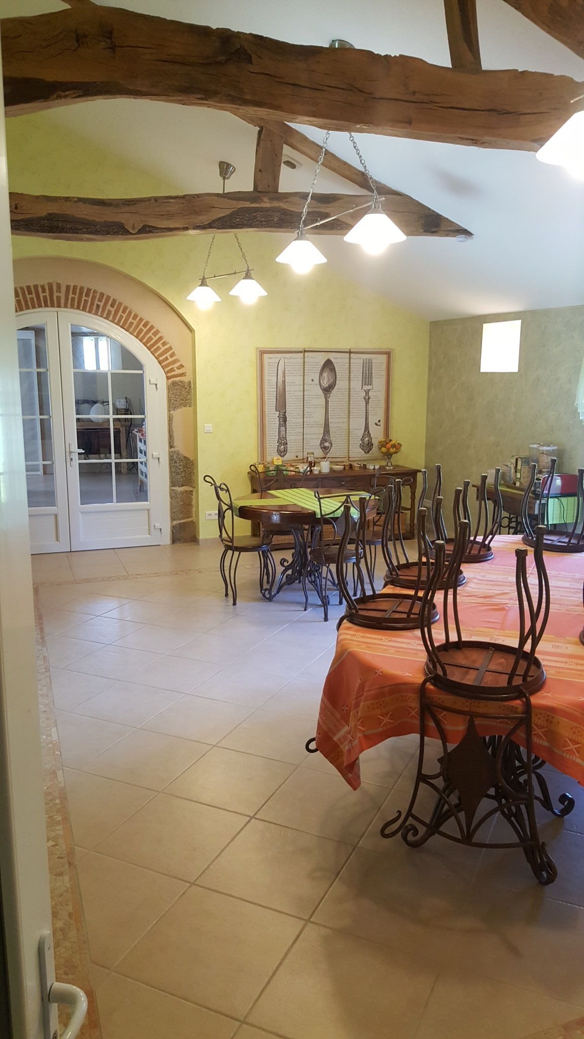 Un comedor con techo alto, vigas de madera a la vista y suelo de baldosas. Hay varias mesas con sillas, un menú colgado en la pared y una puerta que da a otra habitación.