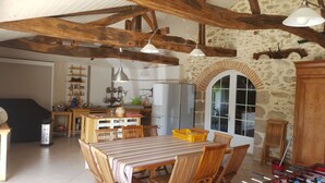 Eine rustikale Küche mit freiliegenden Holzbalken, einem Essbereich mit gestreifter Tischdecke und einer Steinwand mit rundbogenförmigem Fenster.