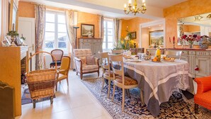 Un coin repas avec une table dressée pour quatre personnes, entourée de chaises. Un lustre est suspendu au-dessus de la table, et une grande fenêtre avec des rideaux se trouve à gauche.