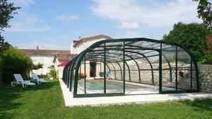 Une piscine couverte, protégée par une structure en verre, entourée d’un mur de pierre et d’une pelouse verdoyante.