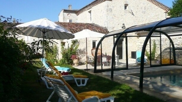 Un espace piscine avec des chaises longues et des parasols, un bâtiment en pierre en arrière-plan et une structure fermée par des parois de verre.