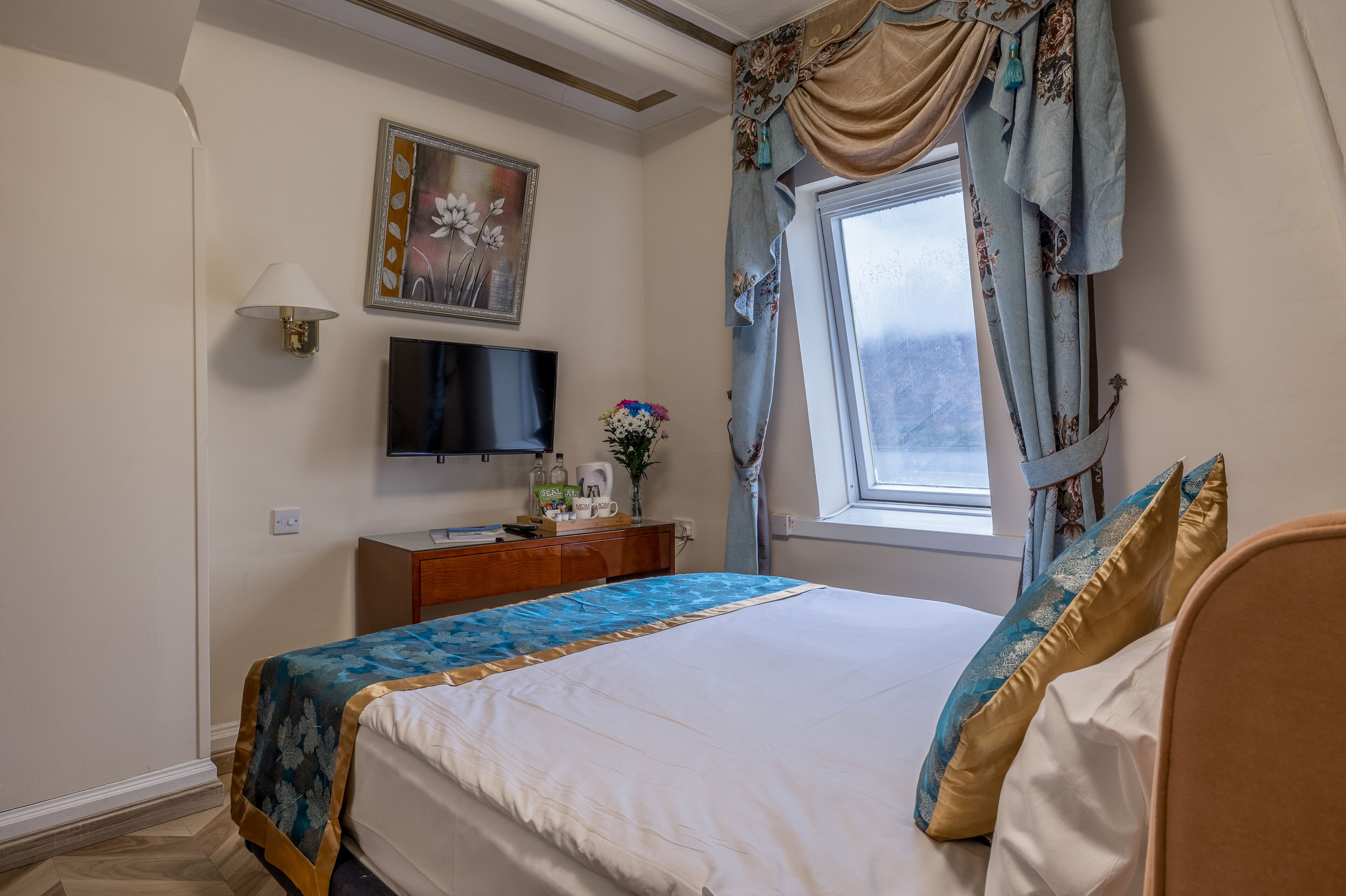 Un dormitorio ordenado con cama, televisor, una mesita con un florero, y una ventana con cortinas.