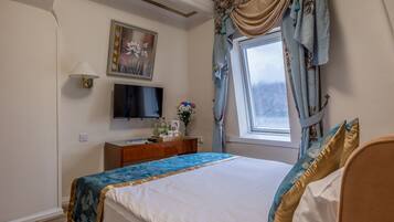 Un dormitorio ordenado con cama, televisor, una mesita con un florero, y una ventana con cortinas.