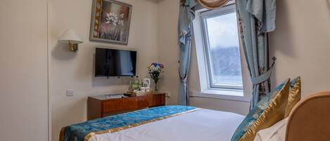 A neatly arranged bedroom with a bed, a television, a small table with a vase of flowers, and a window with curtains.